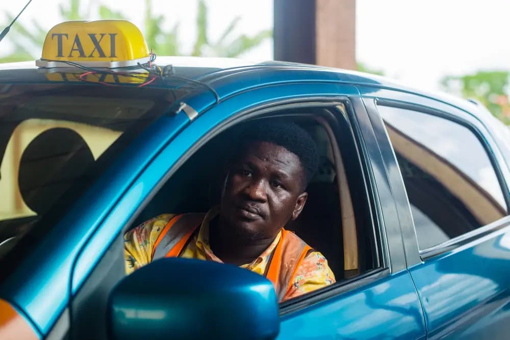 Un chauffeur de taxi en gilet orange, au volant de sa voiture bleue, attend son client