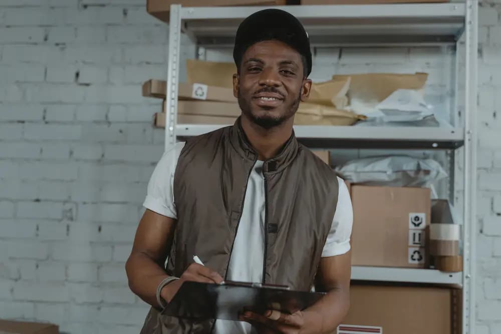 Un magasinier souriant note un inventaire sur sa tablette devant des étagères de colis
